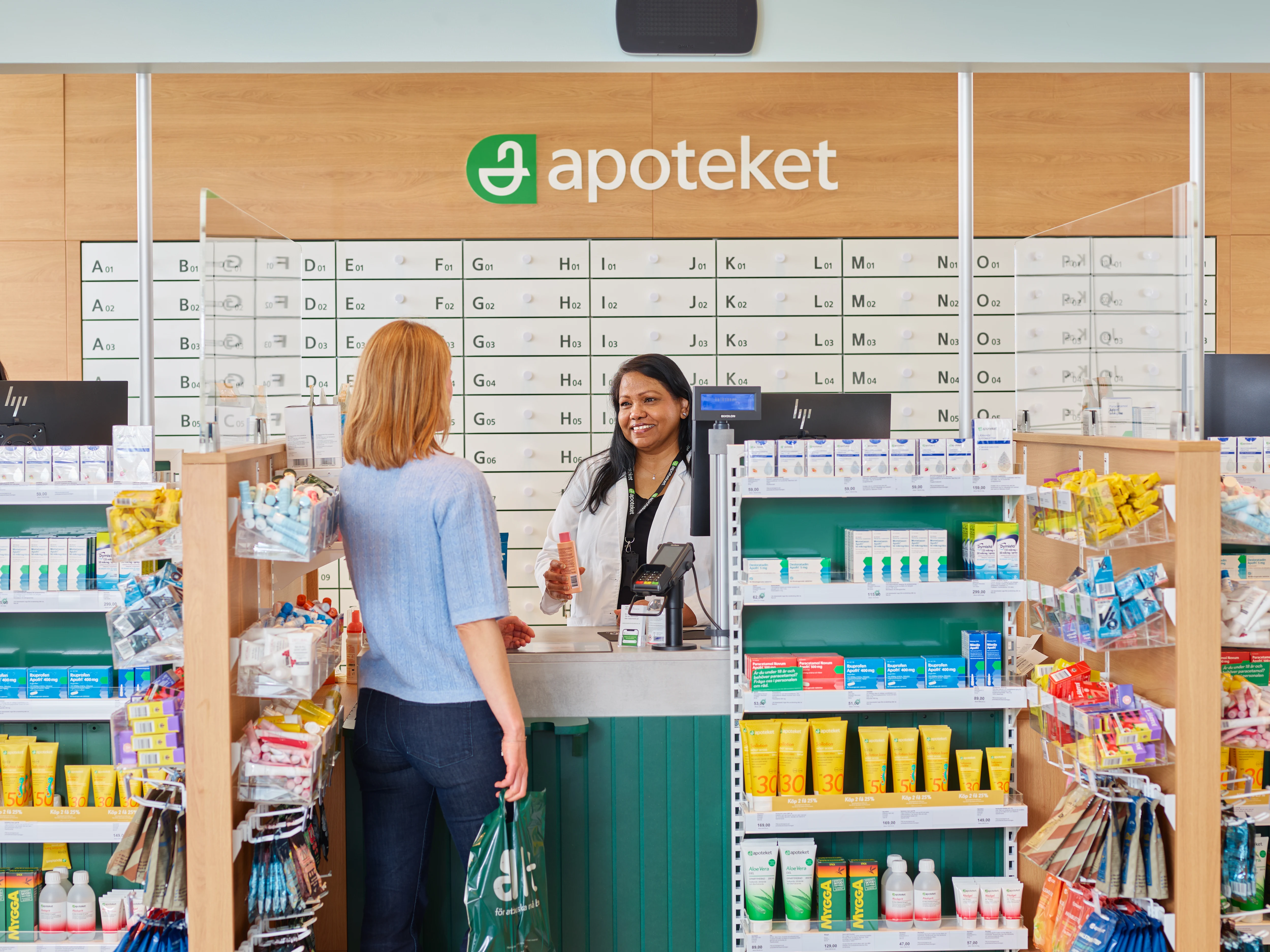En kvinnlig kund står vid disken på ett apotek och pratar med en apotekare. Kunden ser ut att ställa frågor eller få rådgivning om mediciner.