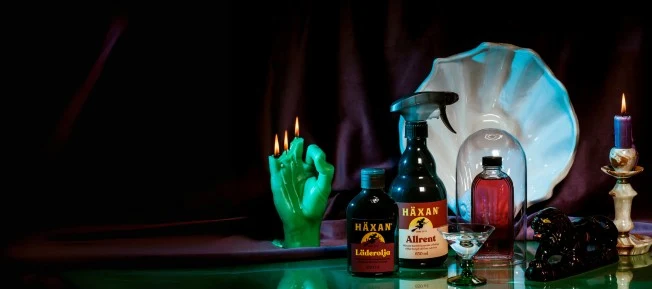 En mystisk uppställning med flaskor, ett grönt handformat ljus, brinnande ljus i olika färger och en vit snäckformad skål mot en mörk bakgrund.