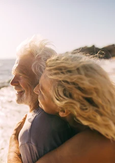 En äldre kvinna och man kramar om varandra på en strand i solen.	