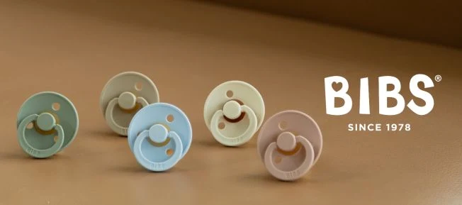 Fem BIBS-nappar i olika pastellfärger (grön, blå, beige, rosa) på en brun yta, med logotypen "BIBS Since 1978" i vitt till höger.